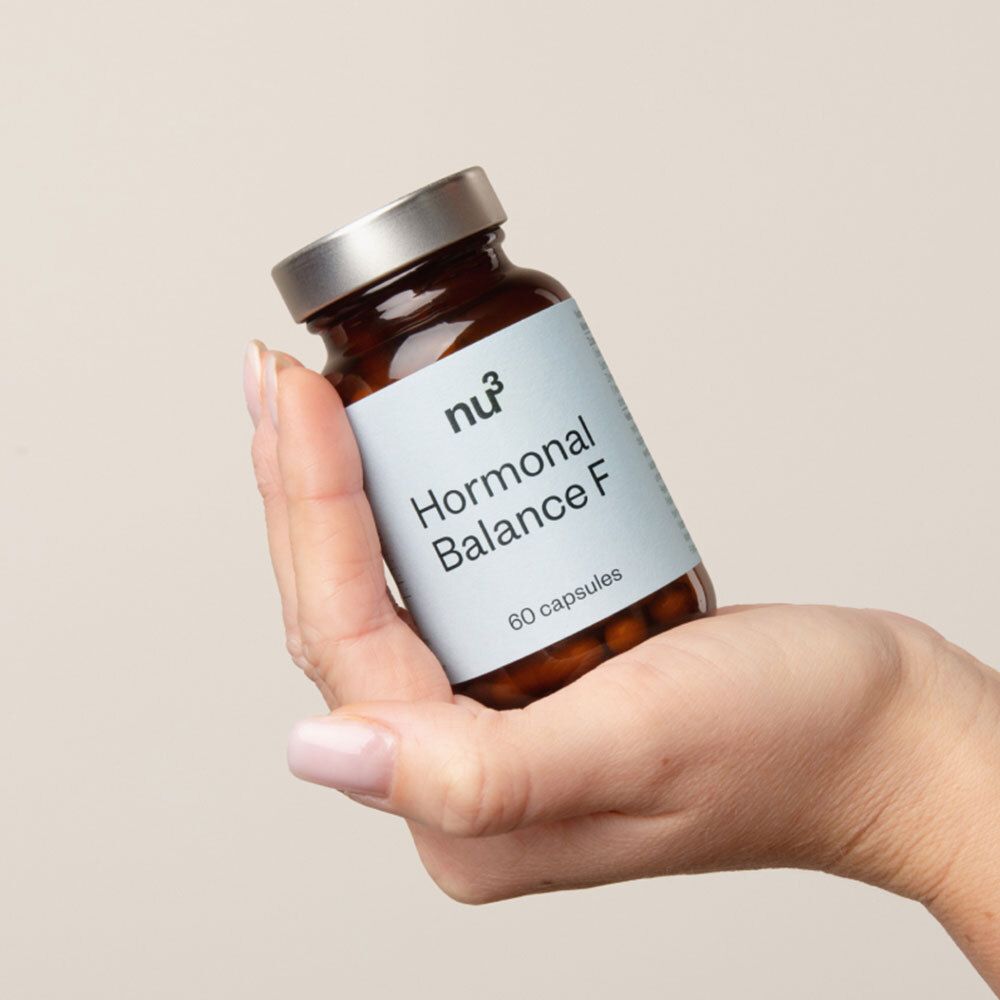 Une main tient un flacon en verre brun avec un couvercle argenté. L'étiquette indique "nu3 Hormonal Balance F" et "60 capsules".