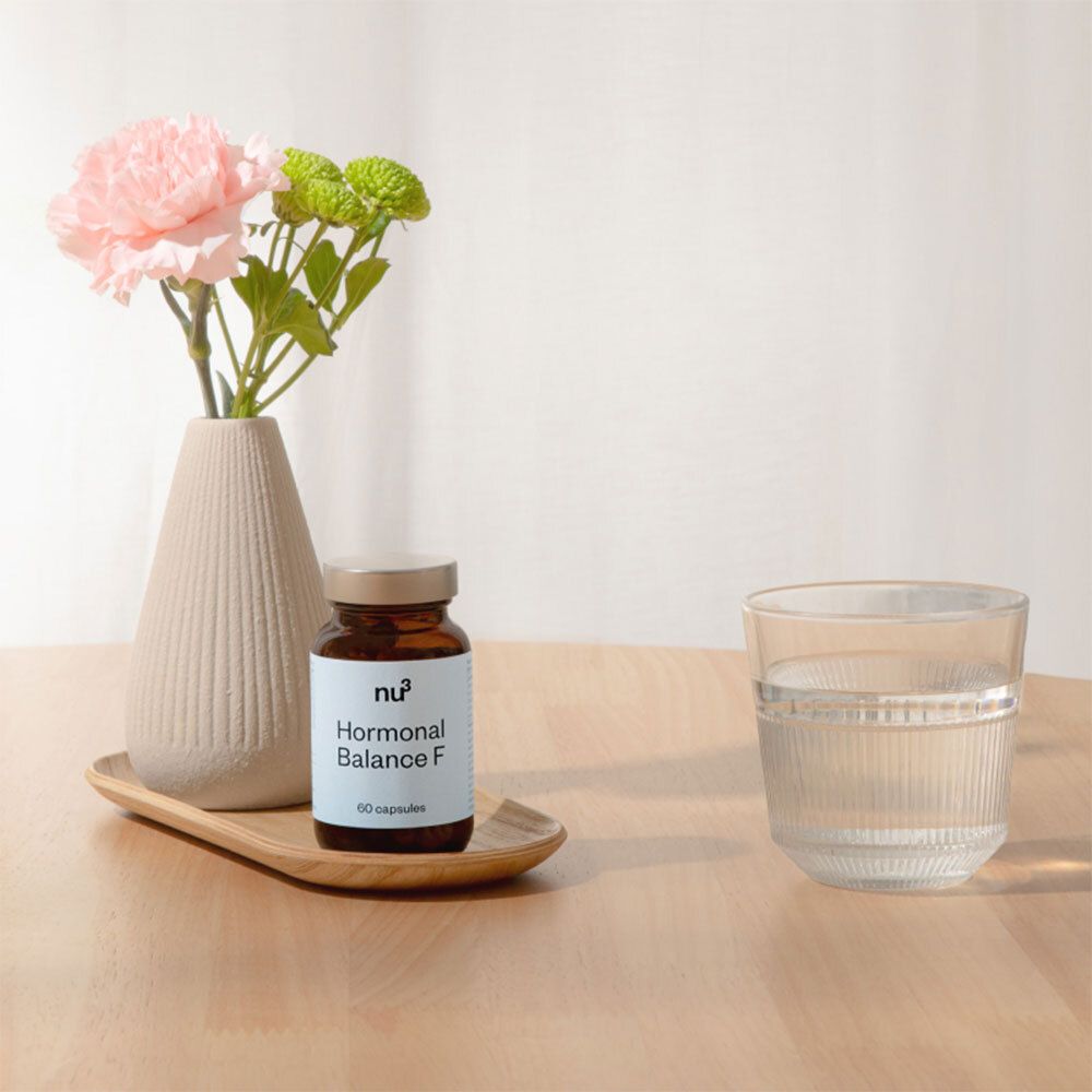 Un flacon en verre brun avec un couvercle argenté est posé sur un plateau en bois, à côté d'un verre d'eau et d'un vase fleuri.