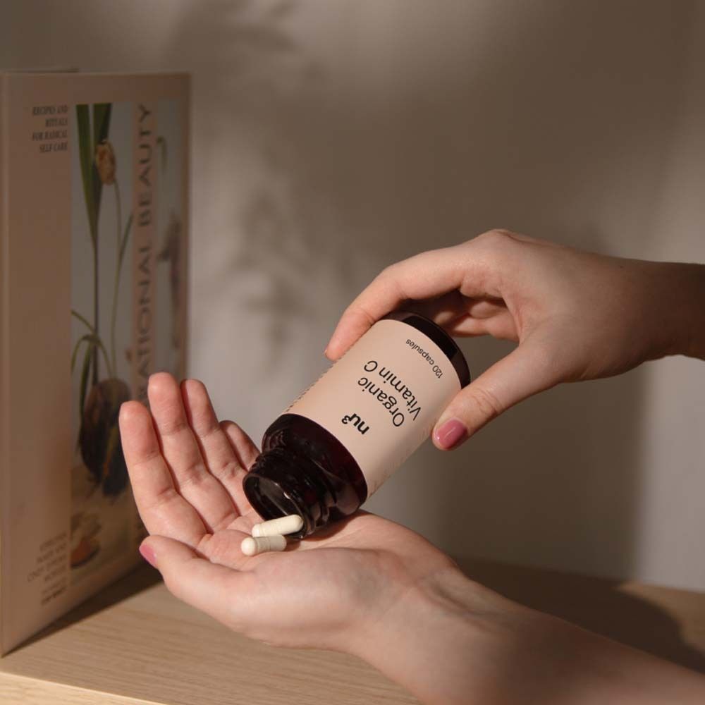 Handen houden een bruine fles "nu3 Organic Vitamin C". Capsules worden in de handpalm gegoten.