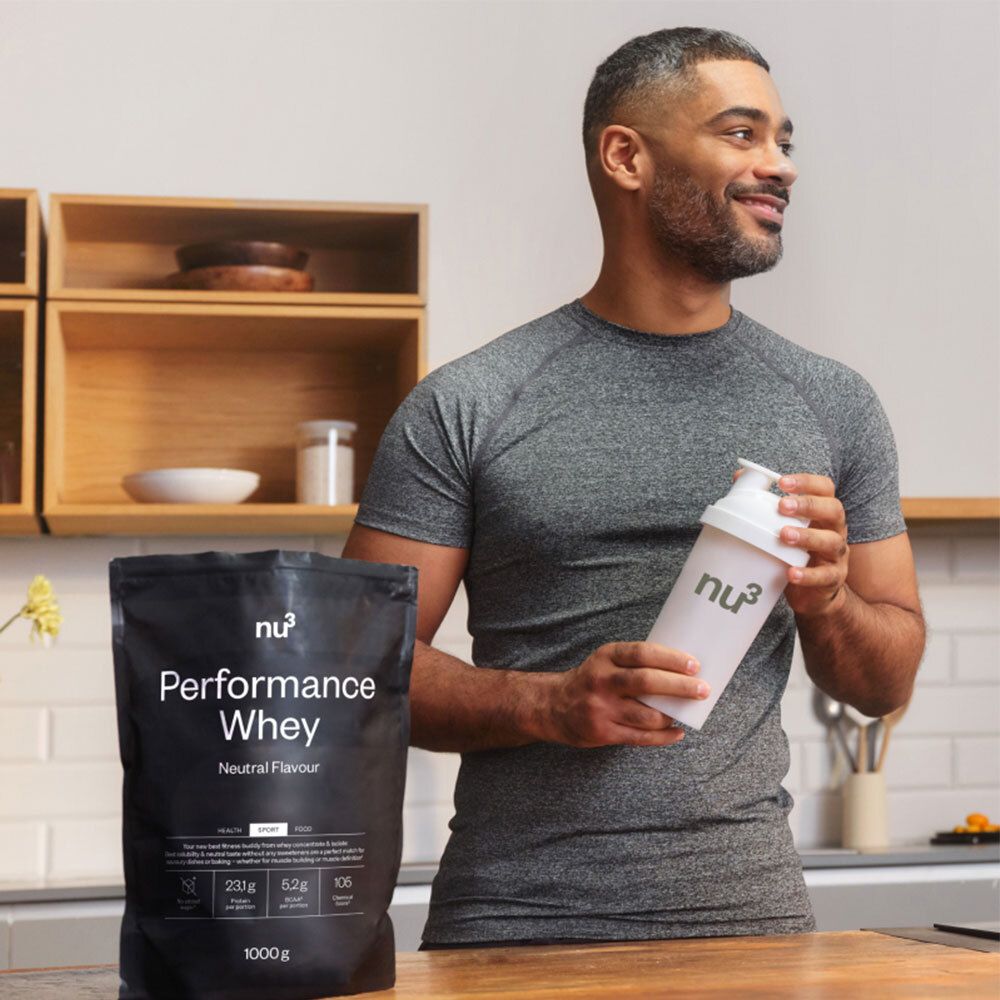 Un homme tient un shaker blanc avec le logo "nu3". À côté de lui : sac noir "Performance Whey". Inscriptions : "Neutral Flavor", informations nutritionnelles. Cuisine en arrière-plan.