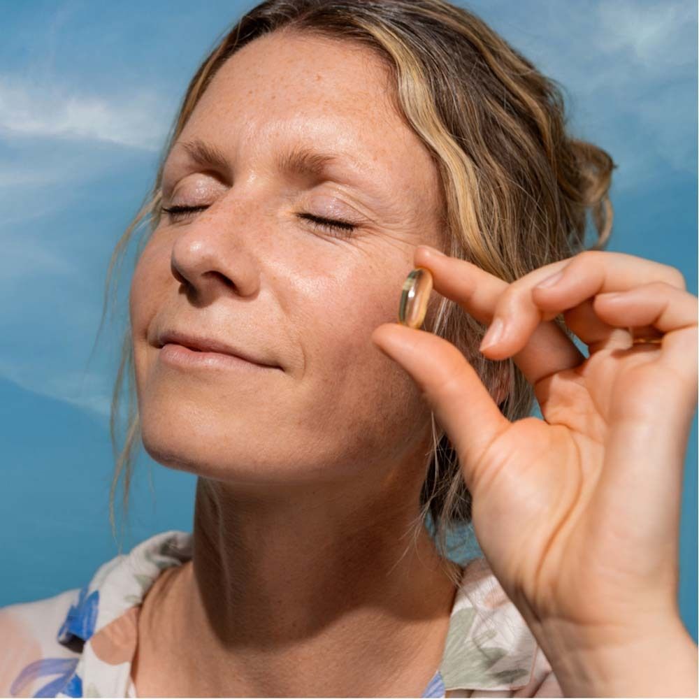 Femme tenant une gélule oméga-3 dorée entre ses doigts. Ciel bleu en arrière-plan.