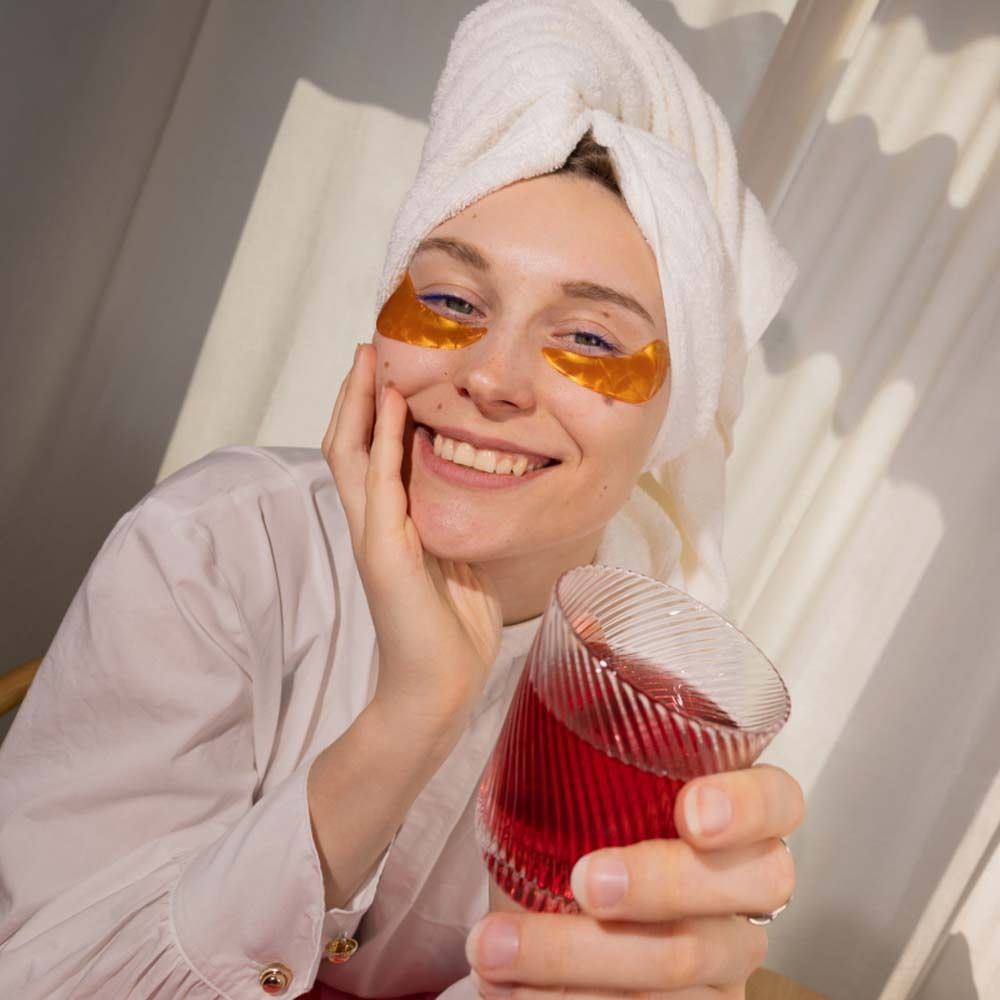Vrouw met oogpads houdt glas met rode drank vast. Op de achtergrond een verpakking.