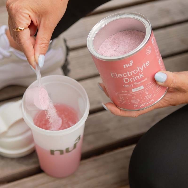 Een hand schept poeder uit een blik nu3 Elektrolyt Drink in een shaker.