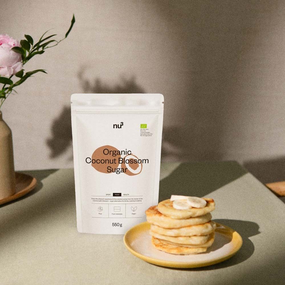 Productverpakking naast pannenkoeken op gele schotel. Bloemen in vaas. nu3 Organic Coconut Blossom Sugar, 550g.