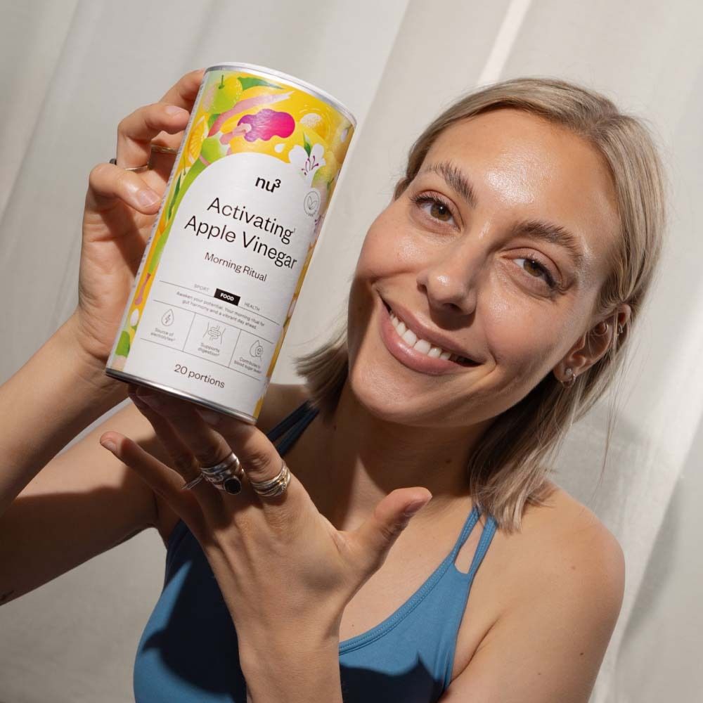 Vrouw houdt doos "nu3 Activating Apple Vinegar" vast. Productnaam en logo zichtbaar. Vrouw lacht.