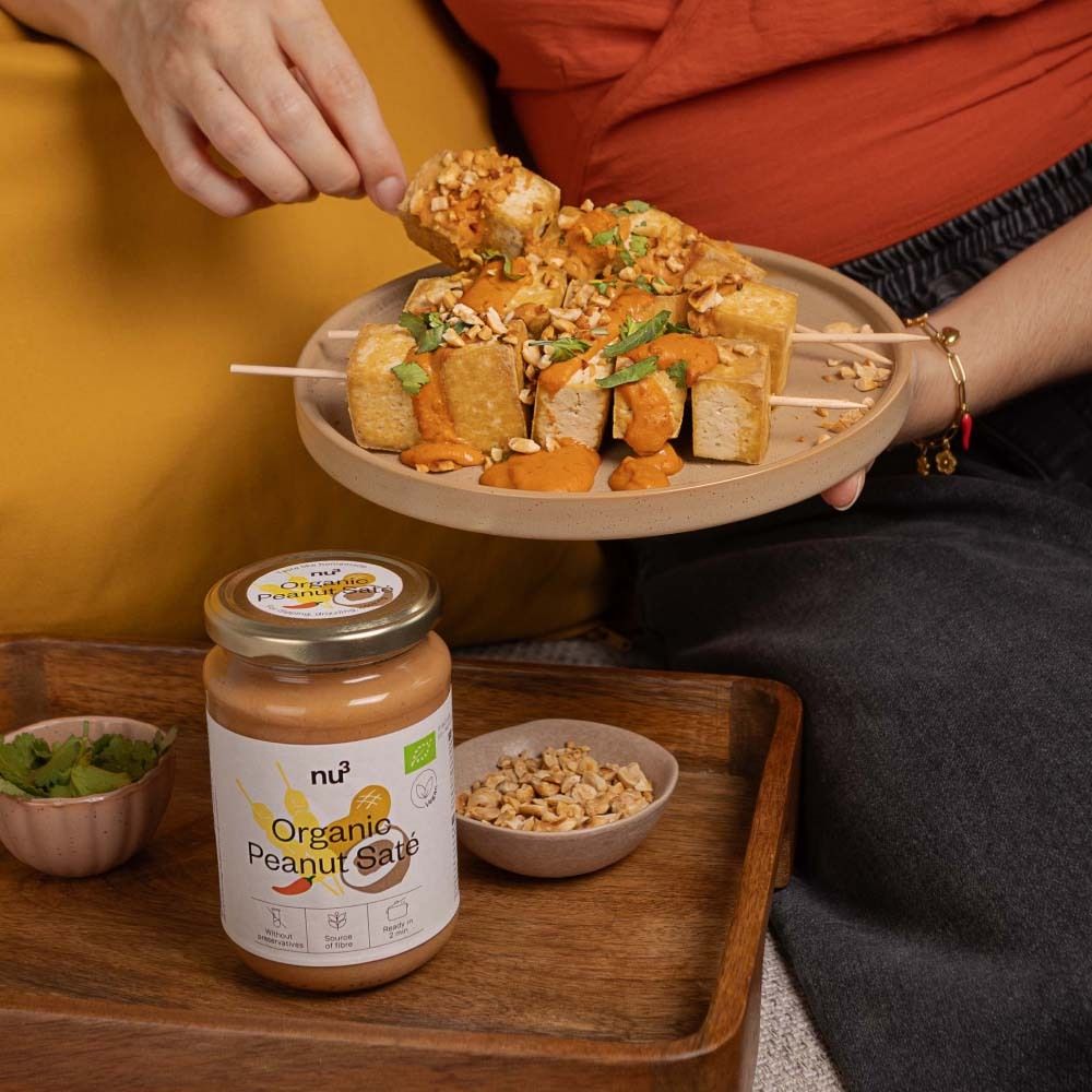 Personne tenant une brochette de tofu et sauce satay aux cacahuètes. À côté, un pot de sauce satay bio nu3 et un bol de cacahuètes.
