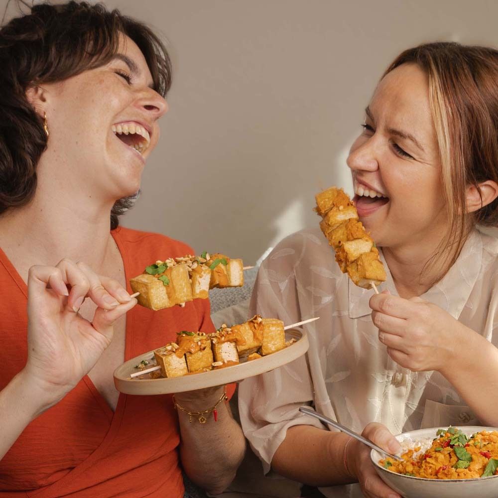 Deux femmes rient, mangent des brochettes de tofu à la sauce satay aux cacahuètes. Sur la table : des bols de nourriture et une assiette de brochettes.