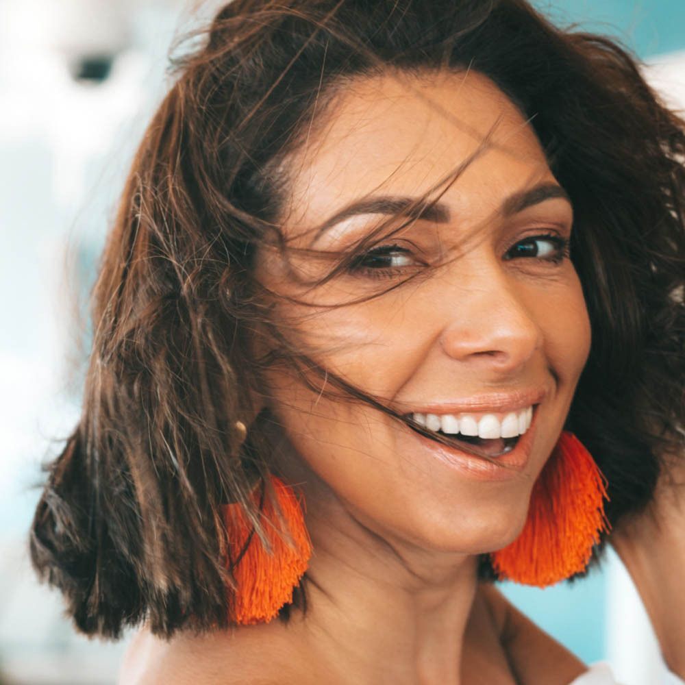 Une femme aux cheveux bruns et aux boucles d'oreilles orange sourit à la caméra.