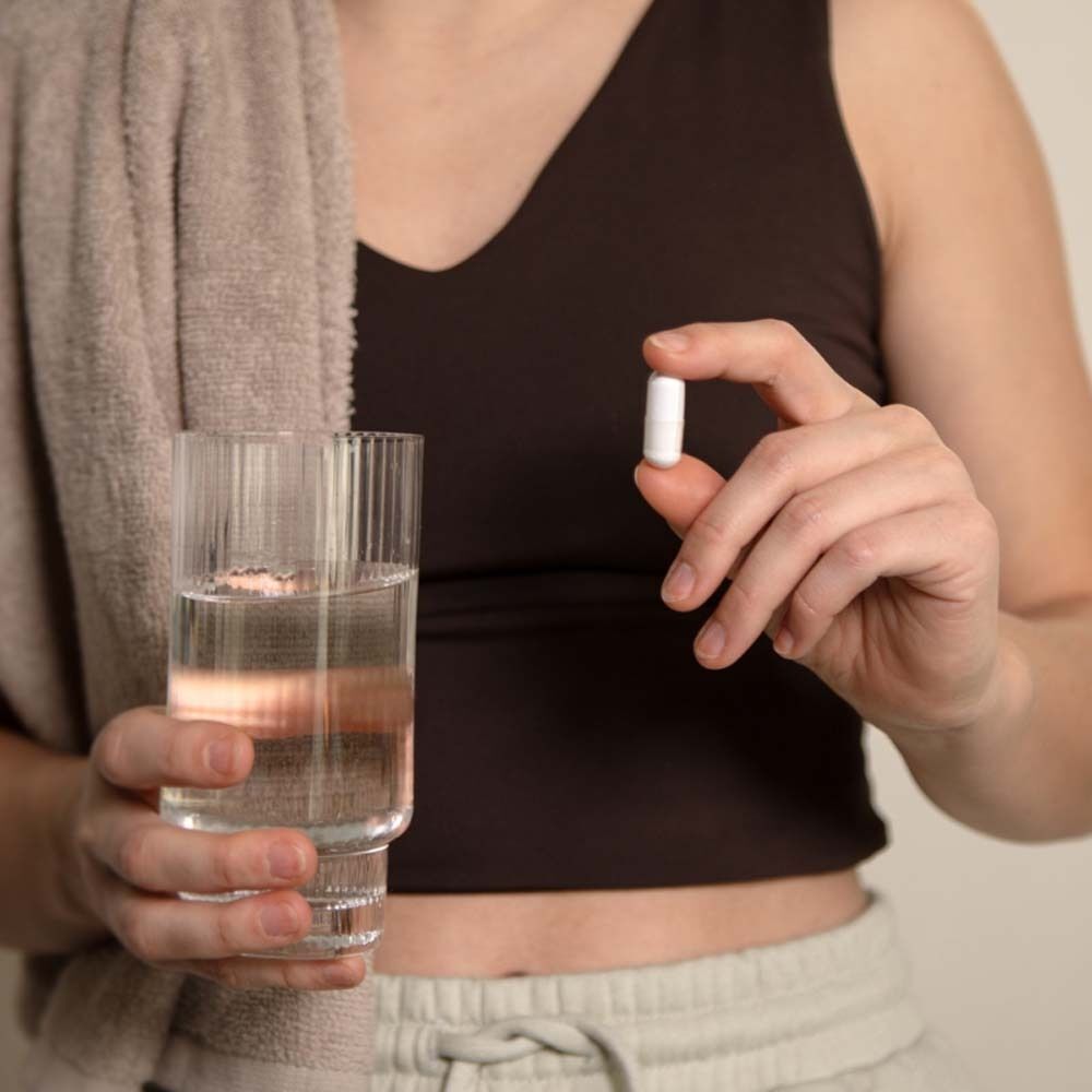 Personne tenant une capsule entre les doigts. À côté, verre d'eau et serviette. La personne porte un haut.