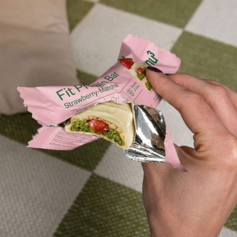 Een hand opent de verpakking van een nu3 Fit proteïnereep, Aardbei-Matcha, met zicht op de reep.