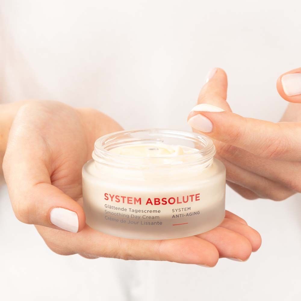 Handen houden een crèmepot vast. Opschrift: SYSTEM ABSOLUTE, Glättende Tagescreme, Smoothing Day Cream, Crème de Jour Lissante.