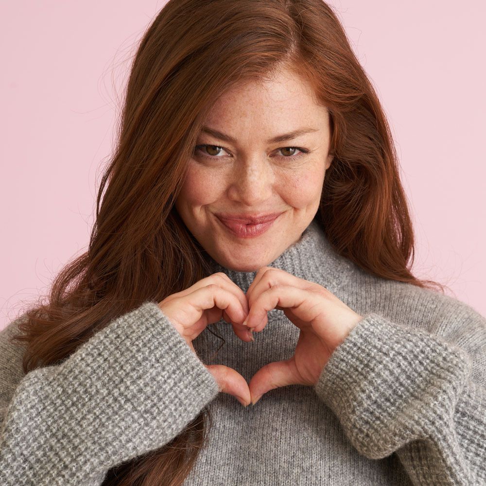 Femme formant un cœur avec ses mains. Pull gris, fond rose.