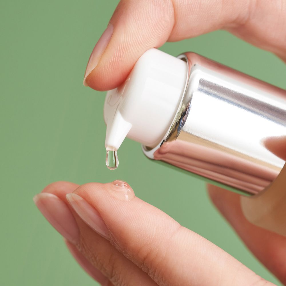 Main pressant le sérum d'un distributeur. Liquide clair qui coule. Fond vert.