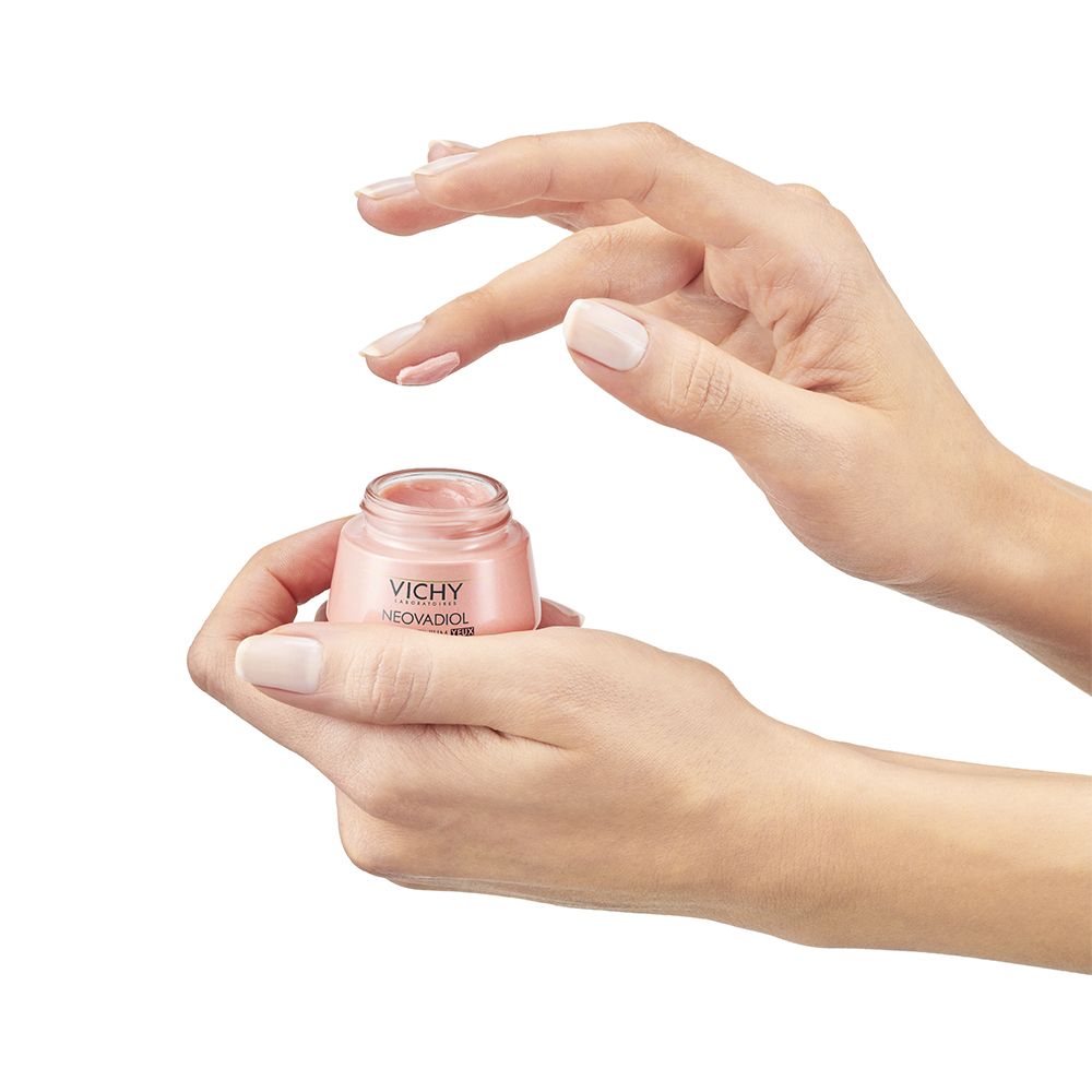Handen met een kleine pot Vichy Neovadiol Rose Platinium oogverzorging, een vingertop raakt de crème aan.