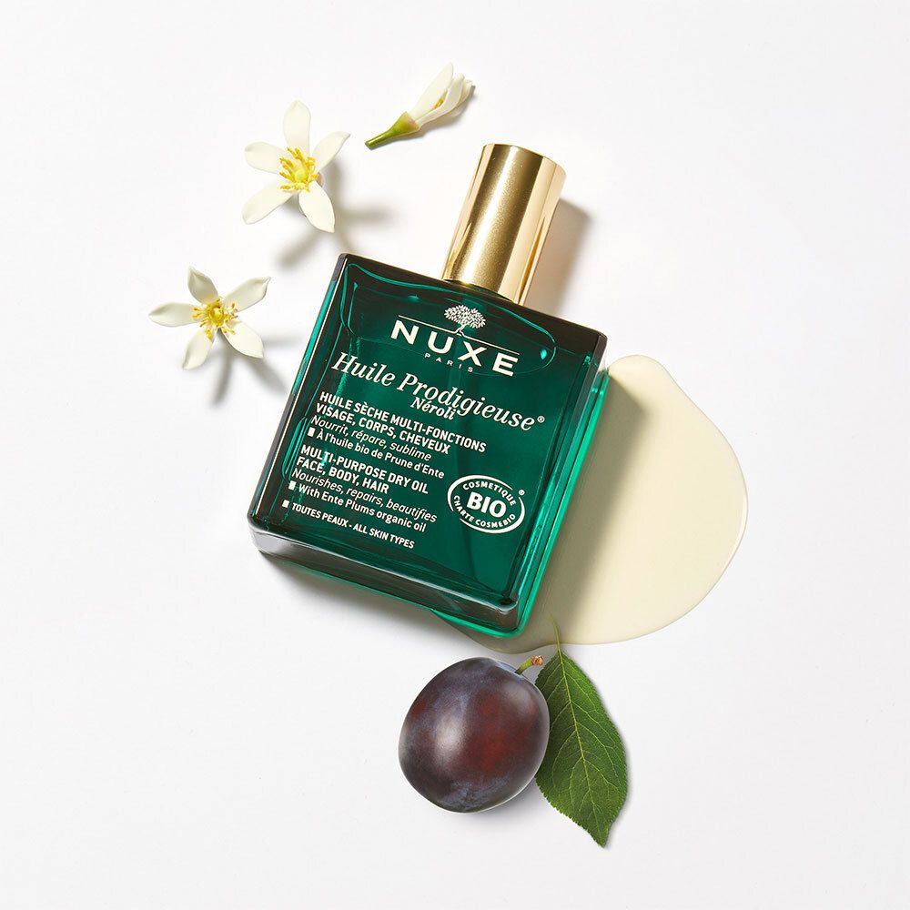 Flacon en verre vert Nuxe Huile Prodigieuse avec bouchon doré, entouré de fleurs et d'une prune.