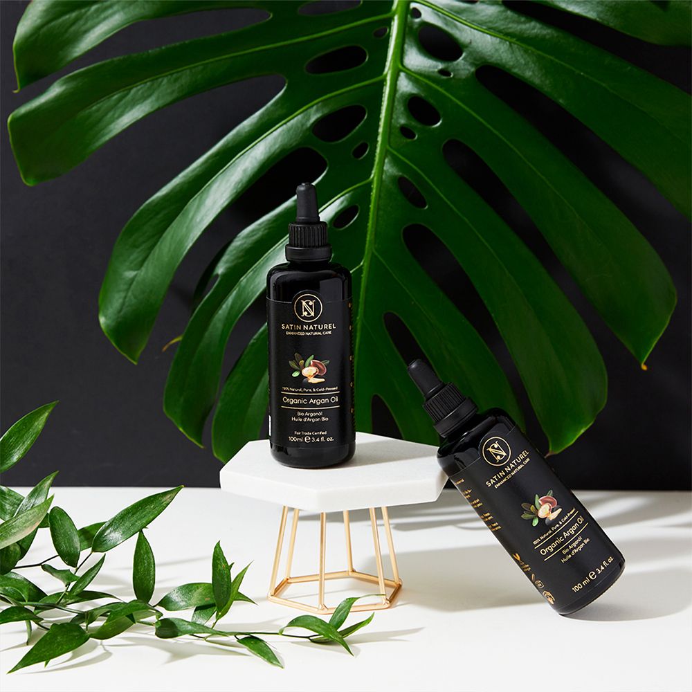 Bouteilles noires d'huile d'argan bio, 50ml et 100ml, avec bouchon compte-gouttes. Feuilles végétales naturelles en arrière-plan.