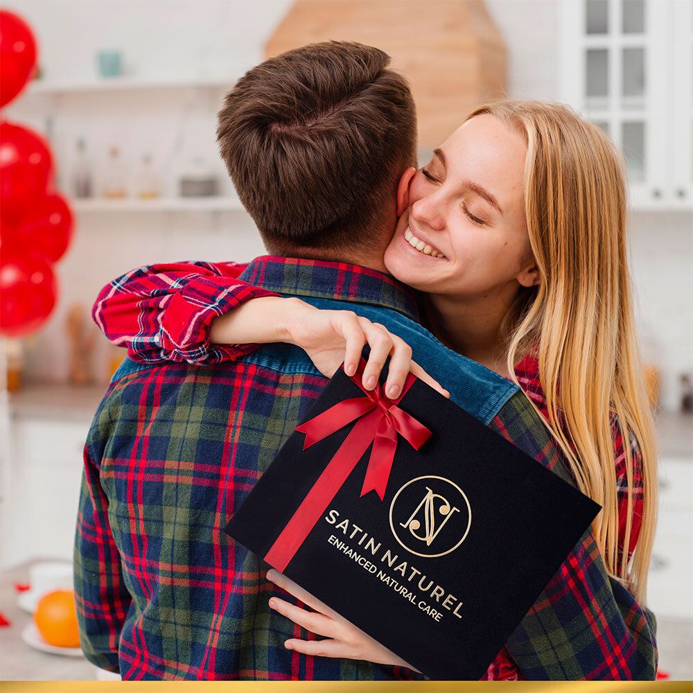 Personne étreignant une personne tenant un coffret cadeau noir avec un ruban rouge. Marque : SATIN NATUREL.
