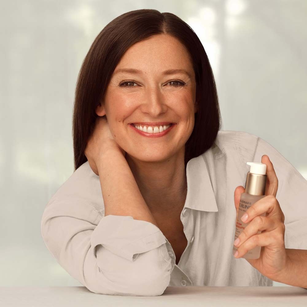 Femme tenant Börlind System Absolute Beauty Fluid. Vêtements clairs. Sourit. Produit en main.