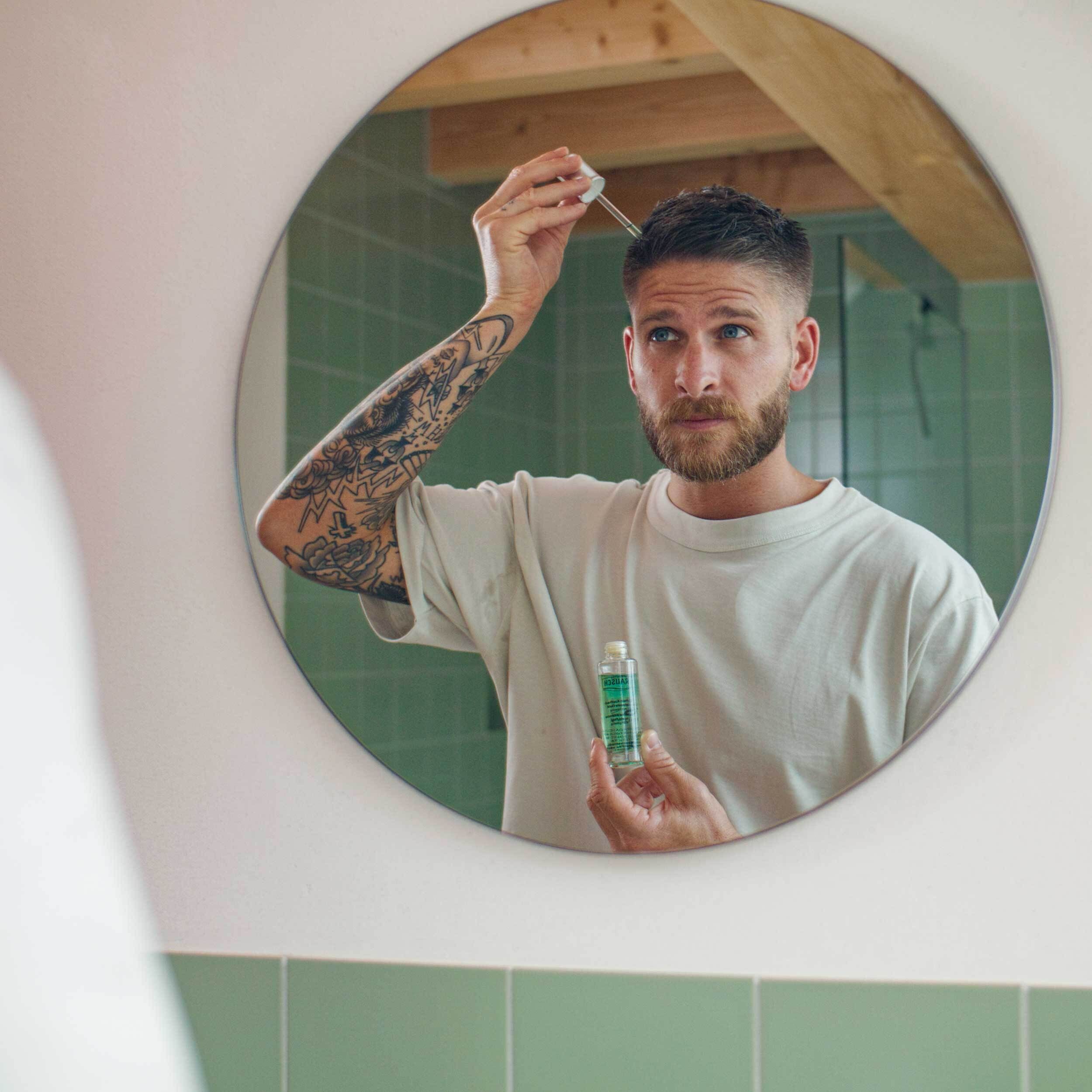 Homme barbu et tatoué applique des gouttes d'une petite bouteille verte avec une pipette sur son cuir chevelu.