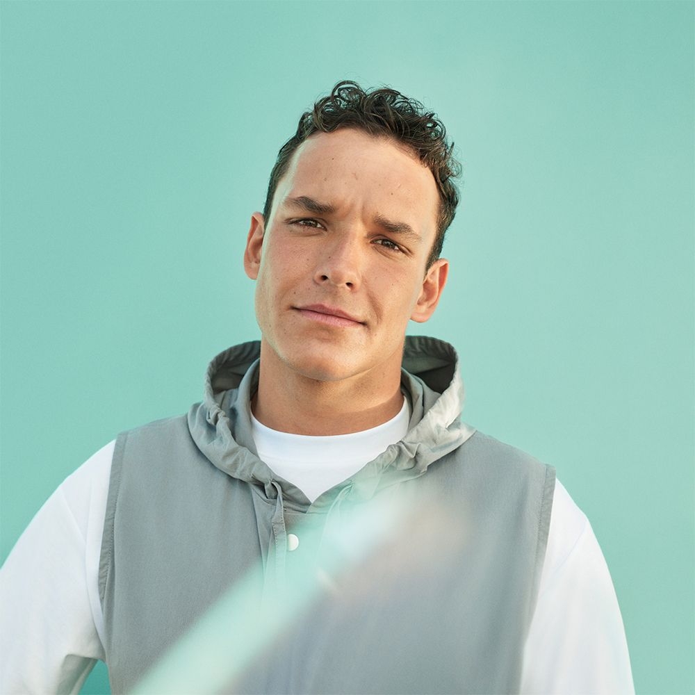 Portrait d'un homme sur fond bleu. Il porte un sweat à capuche gris et un t-shirt blanc.