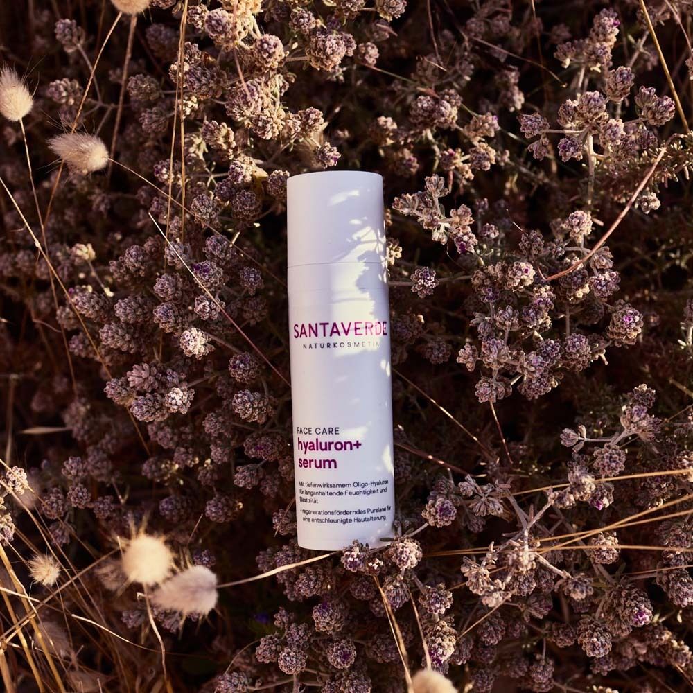 Flacon blanc SANTAVERDE Hyaluron+ Sérum avec applicateur, entouré de fleurs violettes.