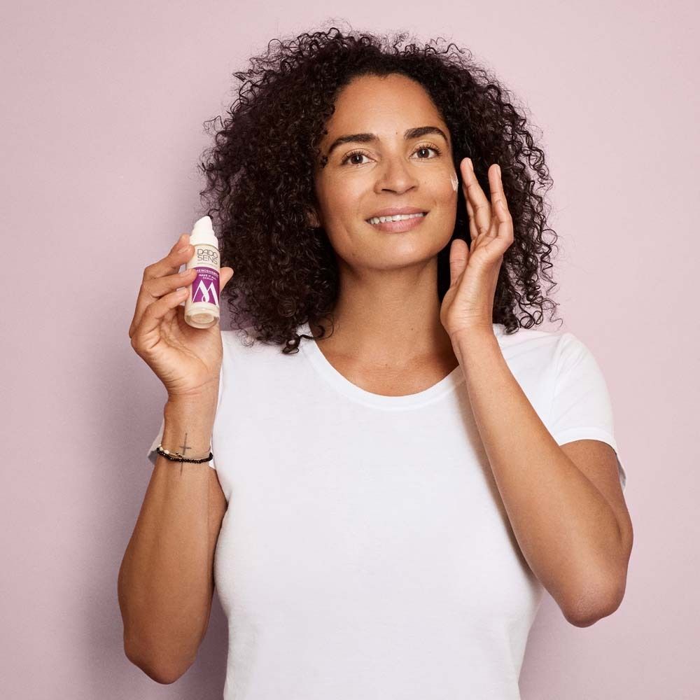 Femme tenant un flacon de sérum Dado Sens Menoenergy. Elle touche son visage. Fond rose.
