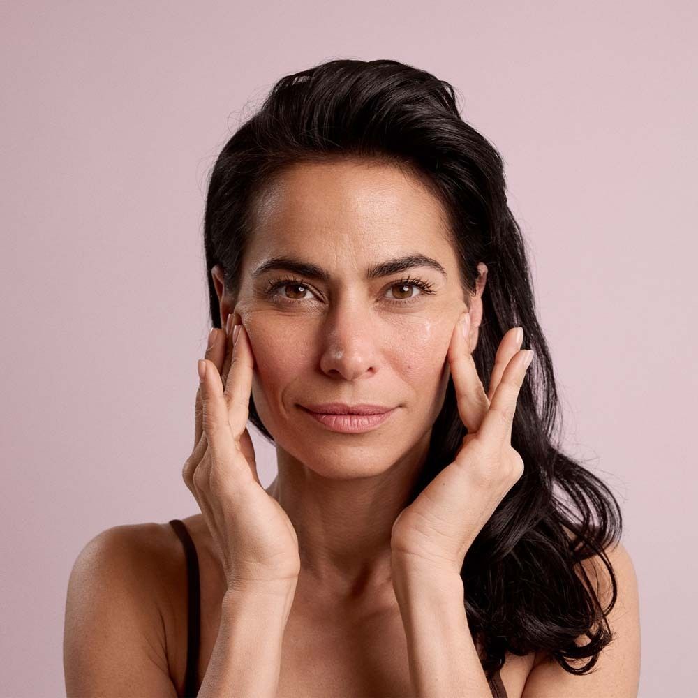 Femme se touche le visage. Elle tient un flacon de sérum Dado Sens Menoenergy. Fond rose.