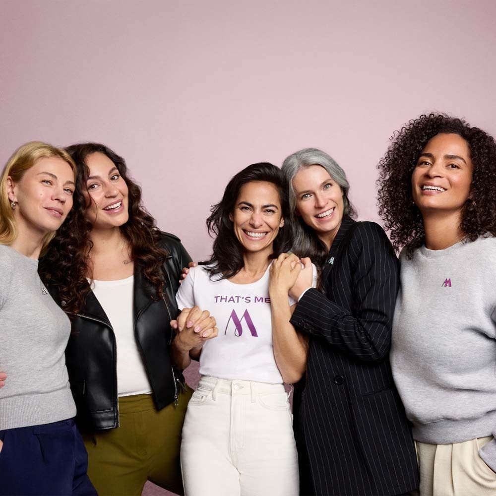Cinq femmes ensemble. Elles portent un t-shirt avec un logo. Fond rose.