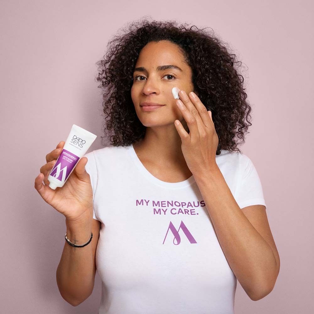 Vrouw met krullen houdt tube vast. Draagt wit T-shirt met logo. Crème op gezicht. Roze achtergrond.