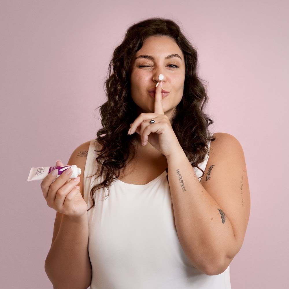 Vrouw met krullen houdt tube vast. Vinger voor mond. Crème op neus. Roze achtergrond.