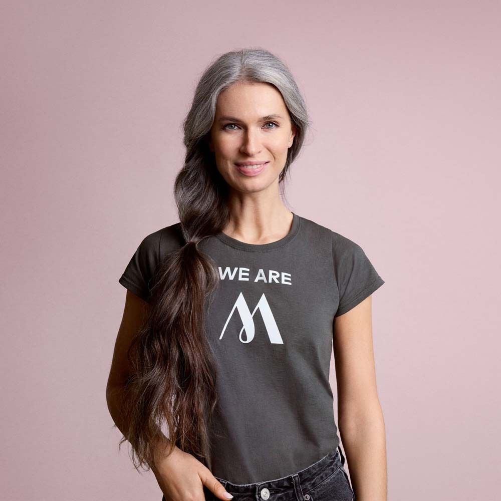 Femme aux cheveux gris et t-shirt gris avec logo blanc. Inscription : WE ARE M.