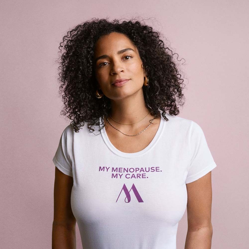 Vrouw in wit T-shirt. Opdruk: My Menopause. My Care. Logo: paarse M. Roze achtergrond.