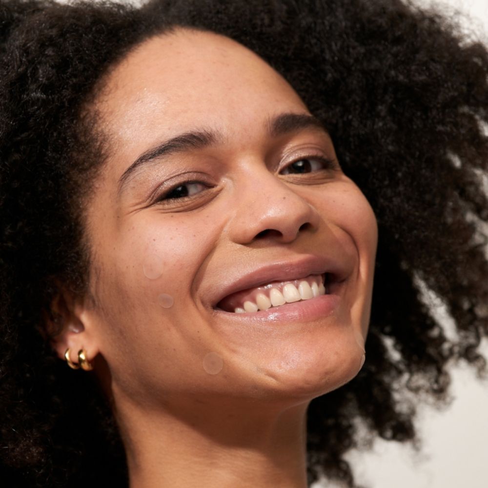 Femme souriante avec des boucles d'oreilles dorées. Deux patchs transparents visibles sur la peau.