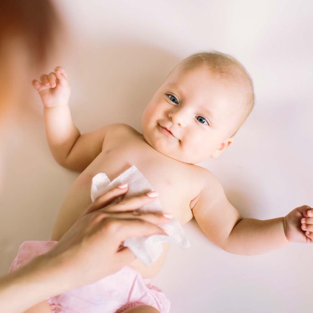 Bébé allongé sur le dos, une main nettoie le haut du corps avec un tissu blanc. Couche rose.
