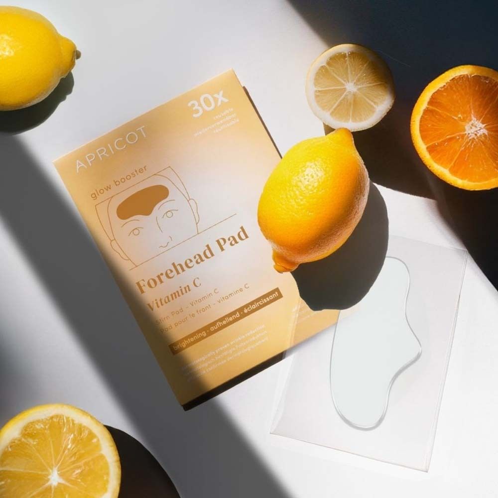 Voorhoofdpad met vitamine C en verpakking. Citroenen en sinaasappel ernaast. Tekst: APRICOT, Forehead Pad, 30x.