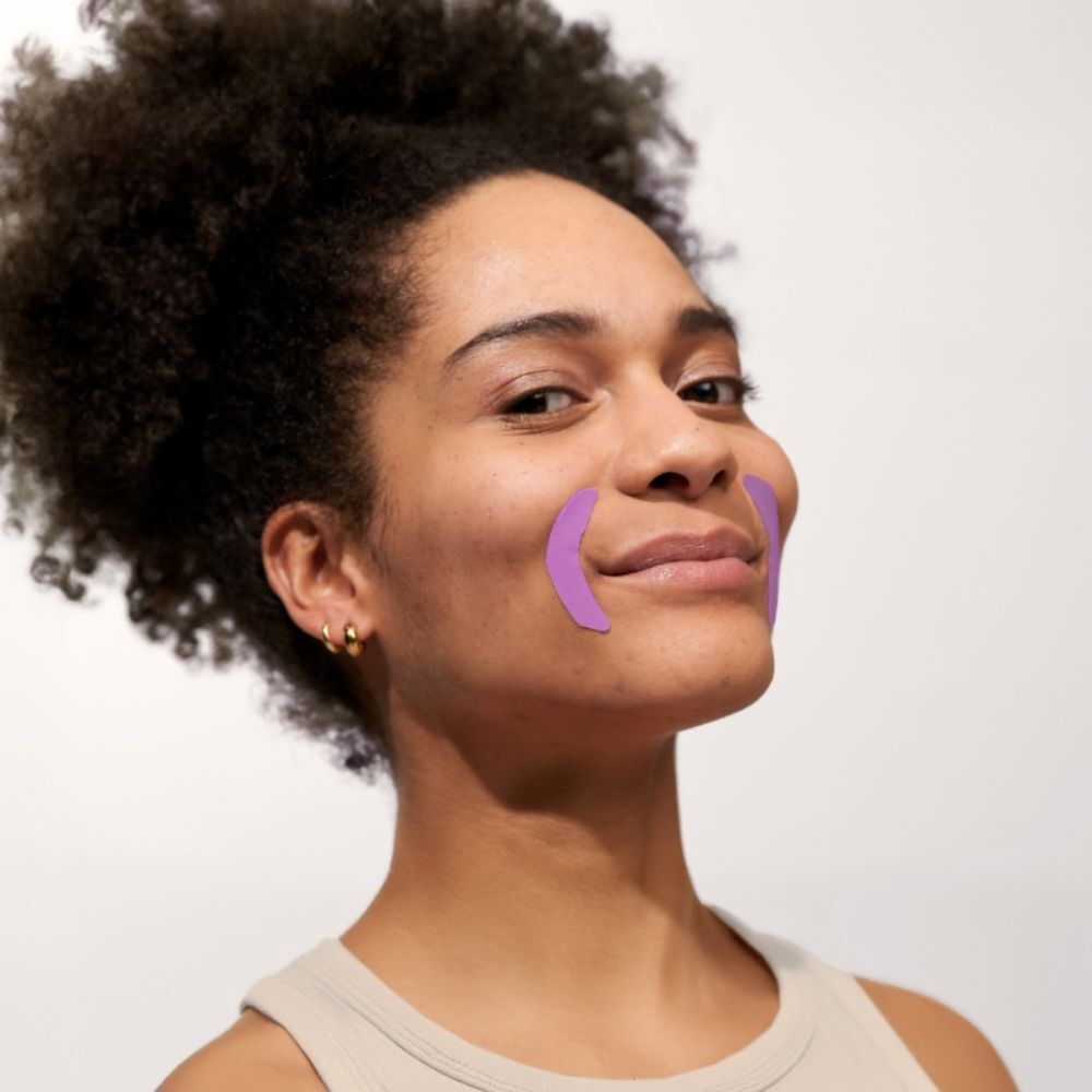 Femme avec deux patchs buccaux violets sur les joues. Elle sourit.