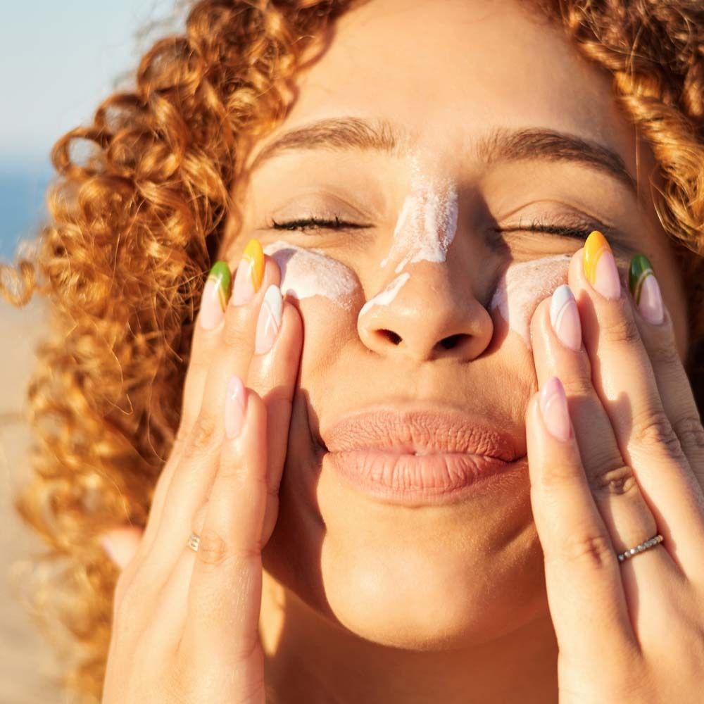 Vrouw brengt zonnecrème aan op haar gezicht. Lichte huid, krullend haar, gekleurde nagels. Close-up.