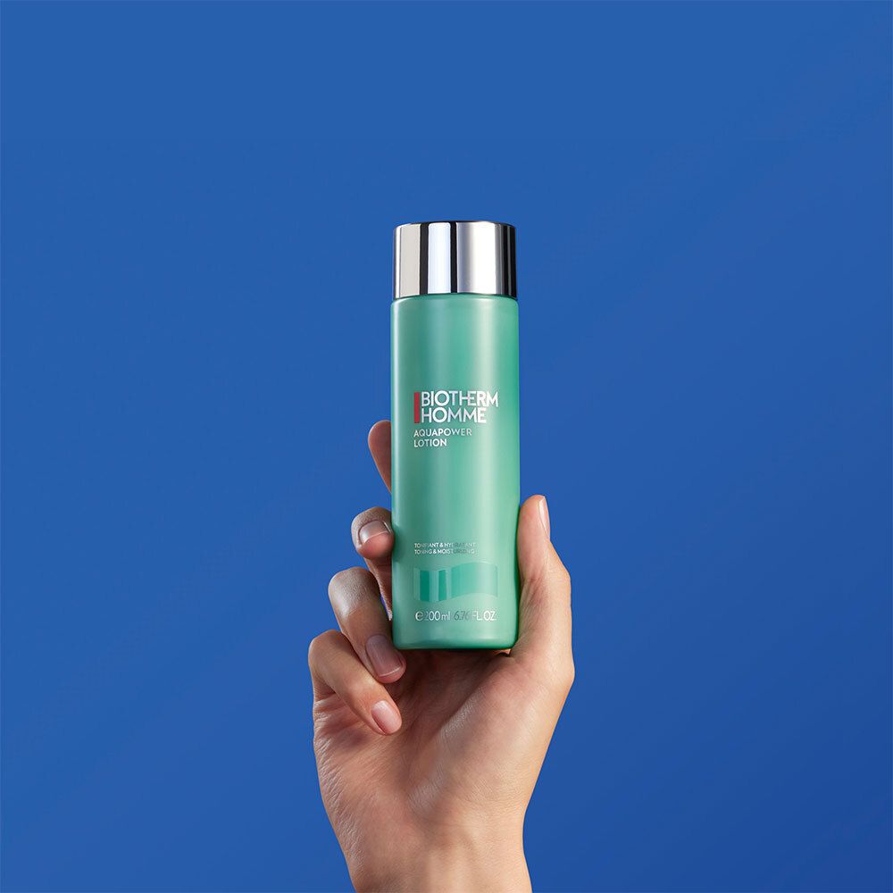 Hand houdt turquoise fles met zilveren dop. Opschrift: Biotherm Homme Aquapower Lotion.
