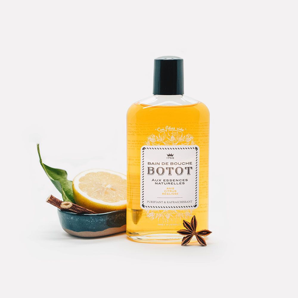 Flacon en verre avec liquide jaune, bouchon noir. Citron, badiane, feuilles et bol à côté. Fond blanc.