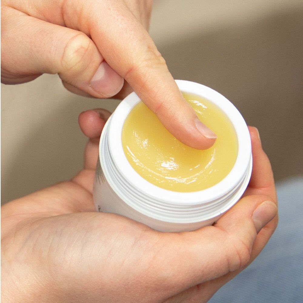 Mains tenant un pot de crème ouvert. Un doigt touche la crème jaune à l'intérieur du pot.