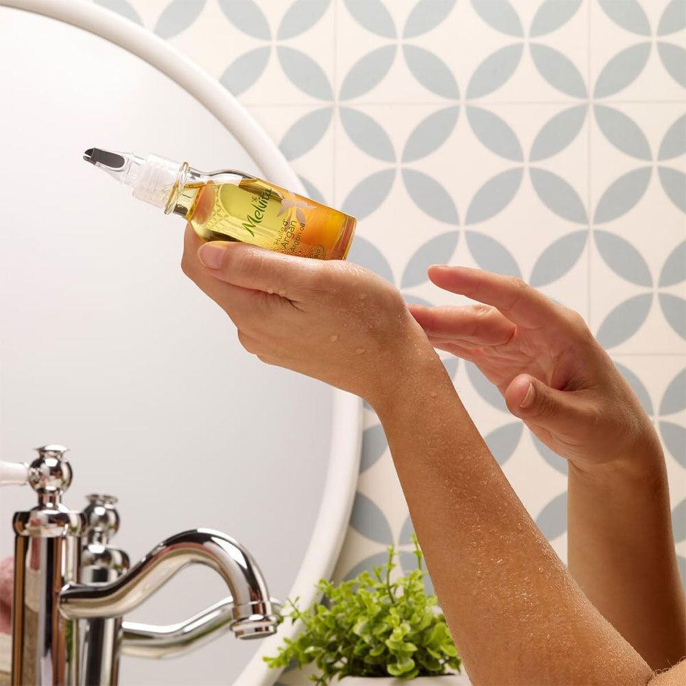 Mains tenant un flacon de Melvita Bio-Mandelöl. Miroir et lavabo en arrière-plan. Flacon avec doseur.