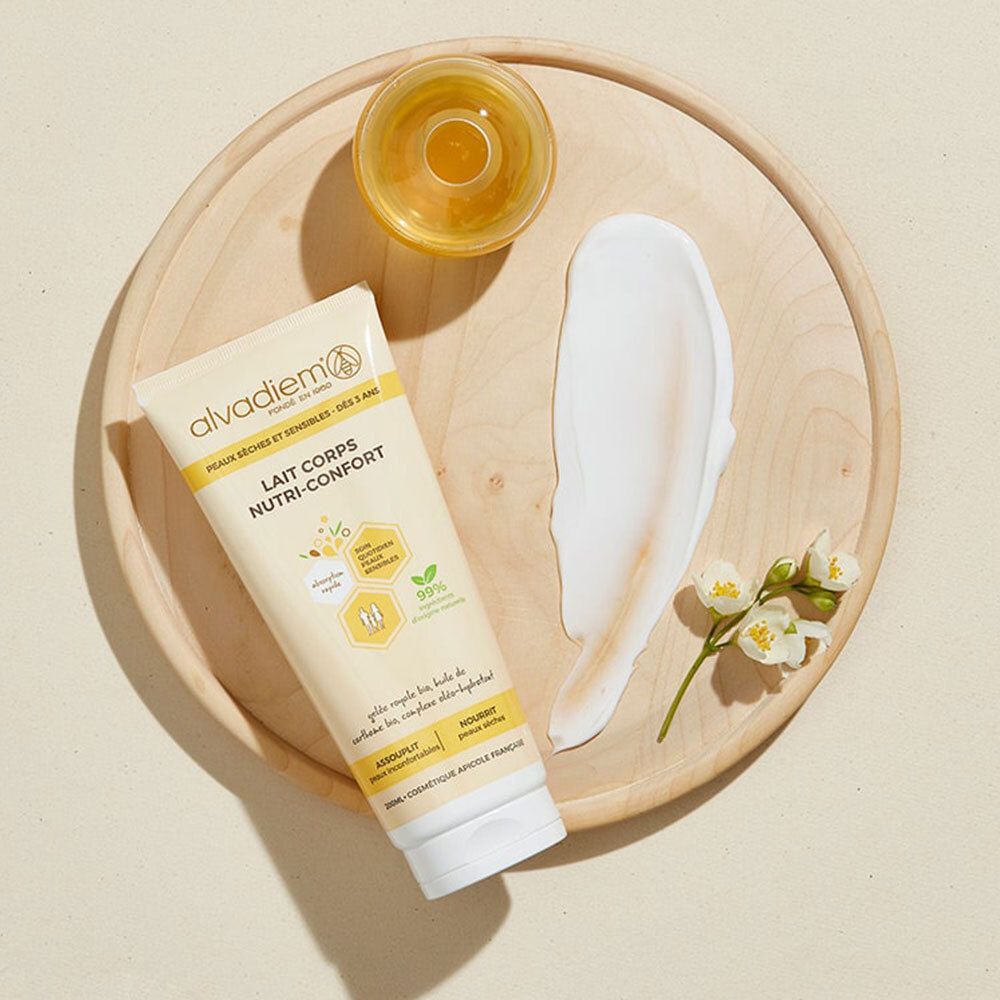 Tube ALVADIEM Lait Corps sur plateau bois, goutte crème, huile et fleurs. Nom du produit et ingrédients sur le tube.