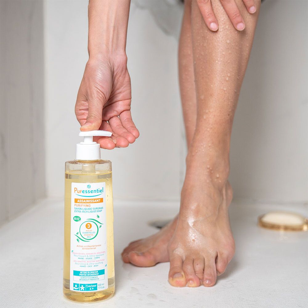 Personne tenant du savon liquide avec pompe. Inscription: ASSAINISSANT, PURIFYING. Personne sous la douche. Pieds et jambes visibles.