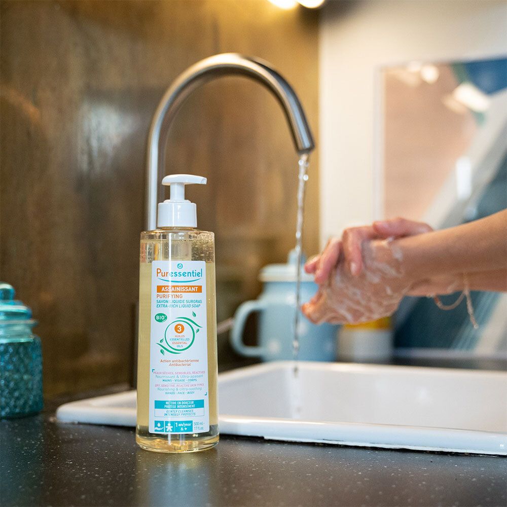 Savon liquide avec pompe près d'un évier. Inscription: ASSAINISSANT, PURIFYING. Mains lavées. Robinet.