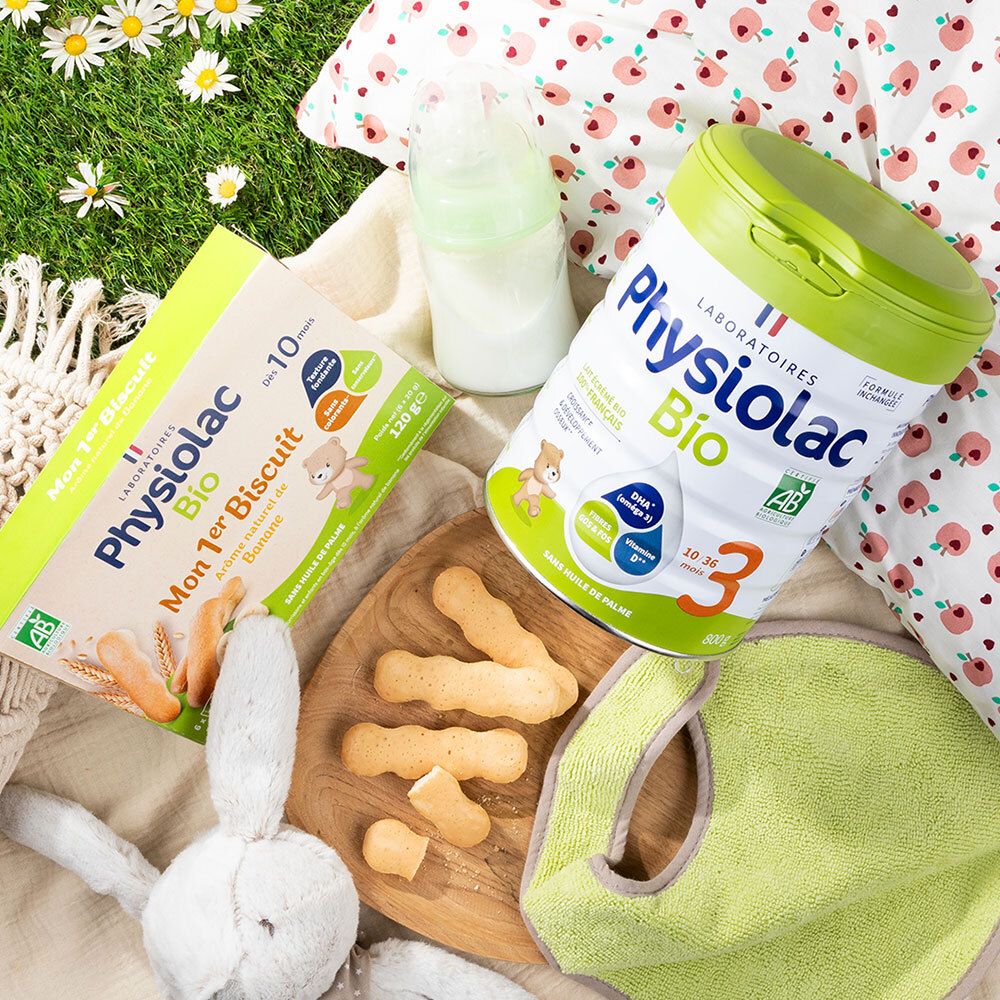 Scène met Physiolac Bio producten. Mon 1er Biscuit Banane, melkpoeder, fles, slabbetje en speelgoed op gras.
