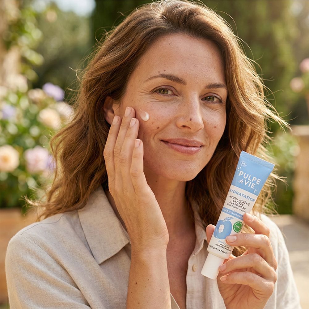 Vrouw houdt tube met productnaam vast. Ze brengt crème aan op haar gezicht. Achtergrond: tuin met bloemen.