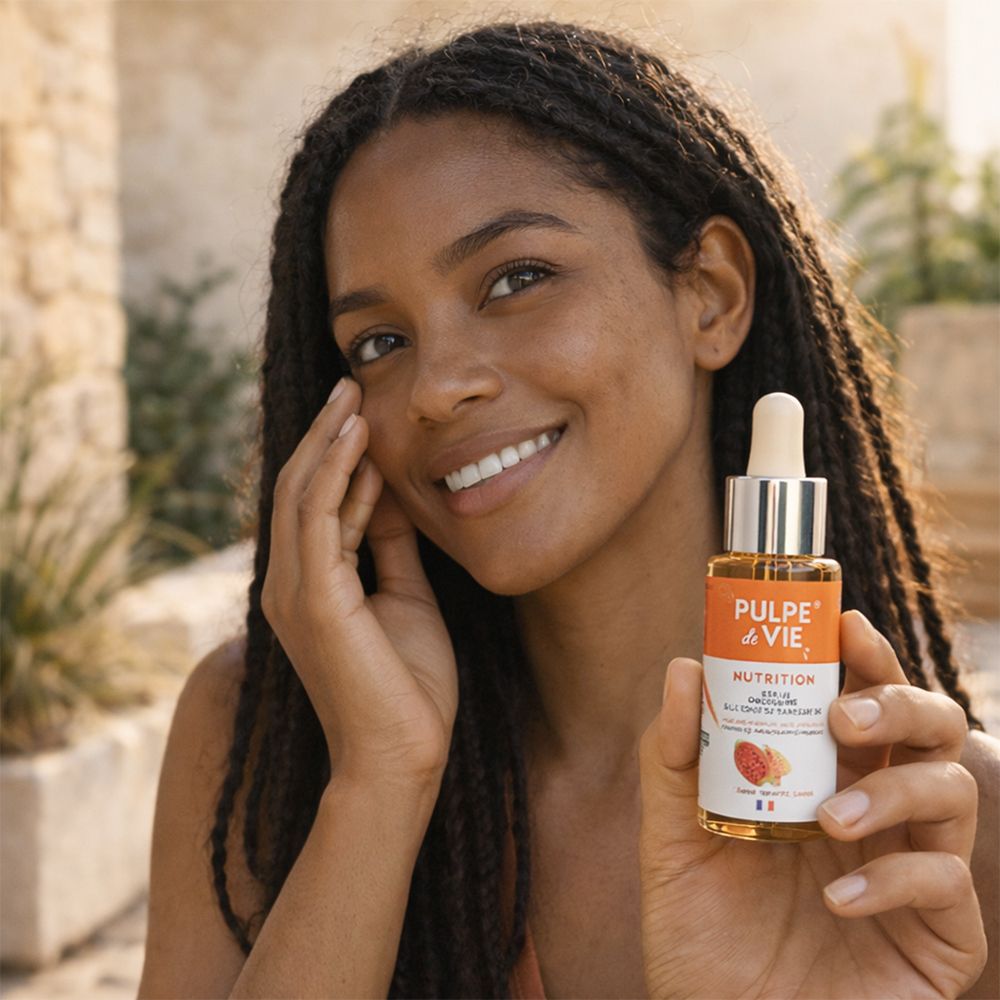 Vrouw houdt serumfles vast. Opschrift: PULPE de VIE, NUTRITION. Gezichtsserum. Vrouw lacht. Achtergrond: stenen muur.