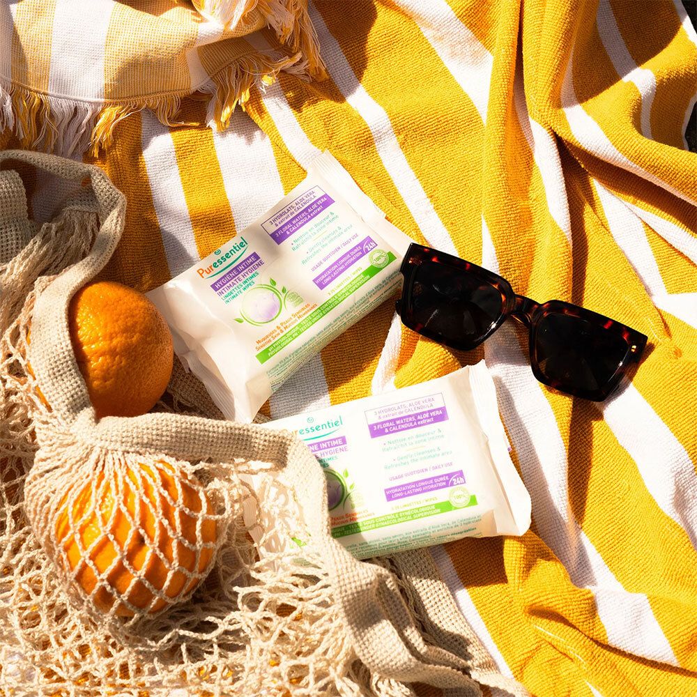 Deux paquets de lingettes intimes sur une serviette rayée jaune et blanche. Texte: HYGIÈNE INTIME. Lunettes de soleil et sac en filet avec orange.