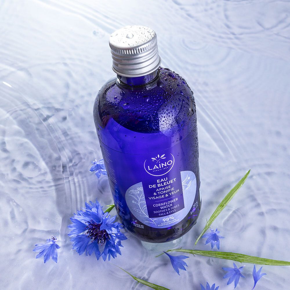 Blauwe fles LAINO Eau de Bleuet in water. Blauwe korenbloemen en groene bladeren omringen de fles. Zilveren dop.