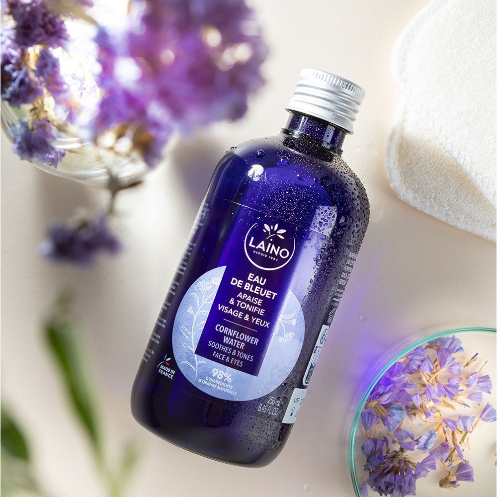 Blauwe fles LAINO Eau de Bleuet, omringd door bloemen en wattenschijfjes. Zilveren dop. Opschrift: Eau de Bleuet, Cornflower Water.